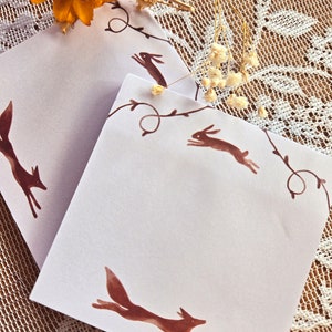 May include: Two white notepads with brown fox and rabbit illustrations on a white lace background. The illustrations are of foxes and rabbits running through a vine.