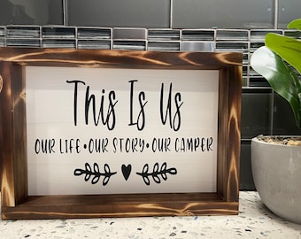 May include: A wooden sign with a white background and black lettering that reads "This Is Us Our Life. Our Story. Our Camper" with a small heart and two sprigs of greenery on either side of the heart.