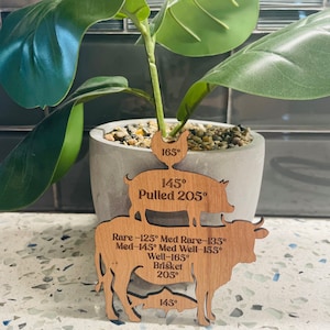 May include: Wooden cutouts of a chicken, pig, and cow with internal temperatures for cooking various meats. The chicken is 165 degrees, the pig is 145 degrees and pulled at 205 degrees, and the cow is 145 degrees for medium rare, 155 degrees for medium well, and 165 degrees for well done. The cow is also labeled as brisket and cooked at 205 degrees.