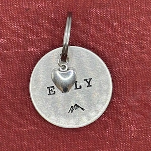 以下が含まれることがあります： 「ELY」の文字が刻印された、銀色の円形ペンダント。上部には小さなハートのチャームがぶら下がり、文字の下には山のグラフィックが刻印されています。ペンダントはシルバーのキーリングに取り付けられています。