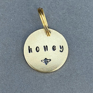 May include: Gold-toned round metal tag with the word "honey" engraved in black lettering and a small bee image. The tag has a gold-toned metal ring attached.