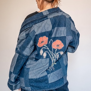 Op de afbeelding: Een gewatteerd spijkerjack met een patchworkontwerp in verschillende blauwtinten en een grote grafische print van oranje klaprozen met stengels en knoppen op de achterkant. Het jack heeft een kraag en lange mouwen met golvend stiksel.