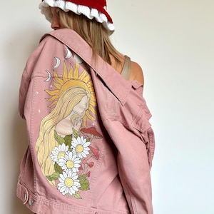May include: A dusty rose denim jacket with a sun and woman illustration on the back. The jacket features a button-down front and pockets. A red mushroom-shaped crochet hat with white trim and white polka dots is worn.