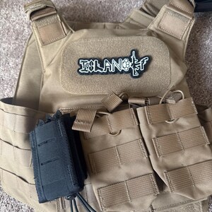 Pode incluir: Colete t&aacute;tico bege com v&aacute;rios bolsos e um porta-carregador preto. O colete apresenta um patch com a palavra "ISLAND" e um gr&aacute;fico de uma arma. Projetado para transportar equipamentos.