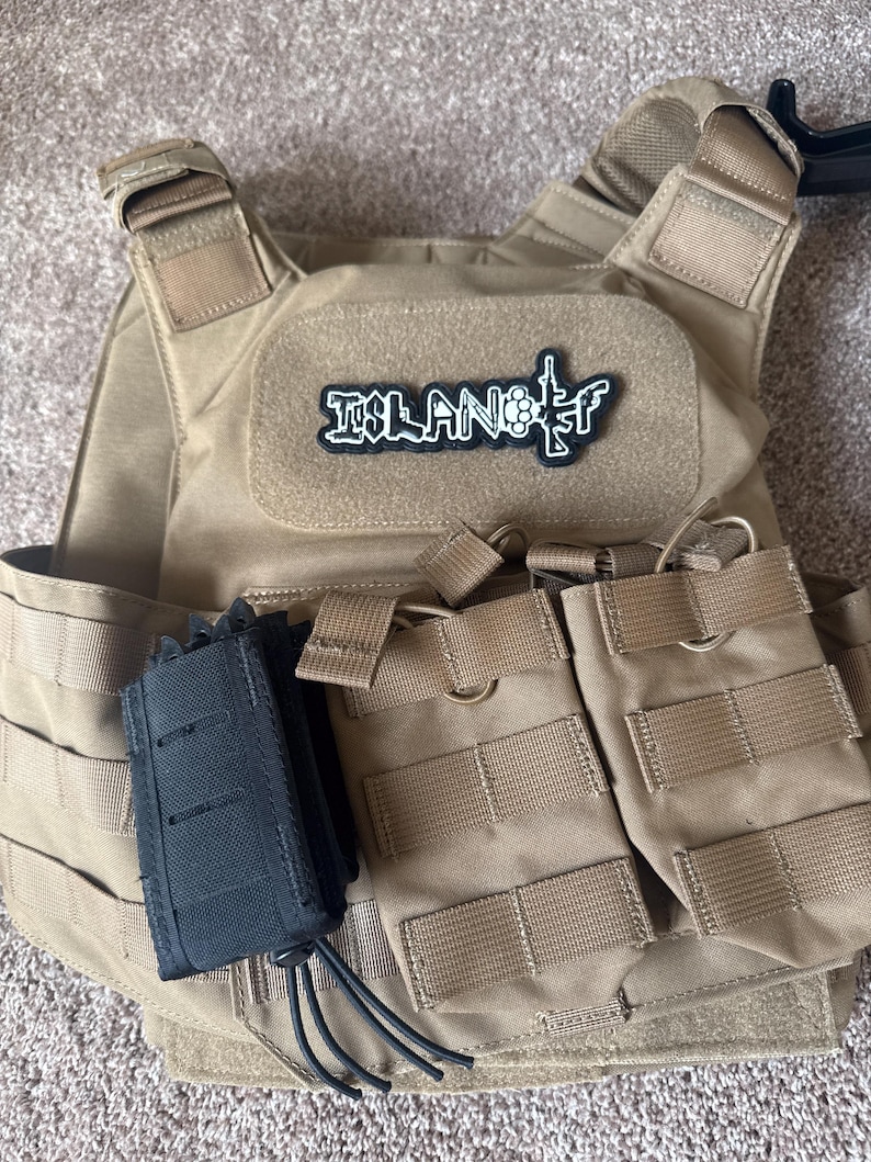 Pode incluir: Colete t&aacute;tico bege com v&aacute;rios bolsos e um porta-carregador preto. O colete apresenta um patch com a palavra "ISLAND" e um gr&aacute;fico de uma arma. Projetado para transportar equipamentos.