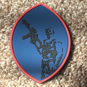 May include: A blue and red patch featuring a black silhouette of a soldier holding a rifle. The patch has a red border and is in an oval shape.