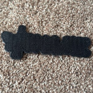 Pode incluir: Um patch preto com a palavra "dreamer" em fonte cursiva. O patch tem uma superf&iacute;cie texturizada e uma borda ondulada. O patch est&aacute; sobre um tapete castanho.