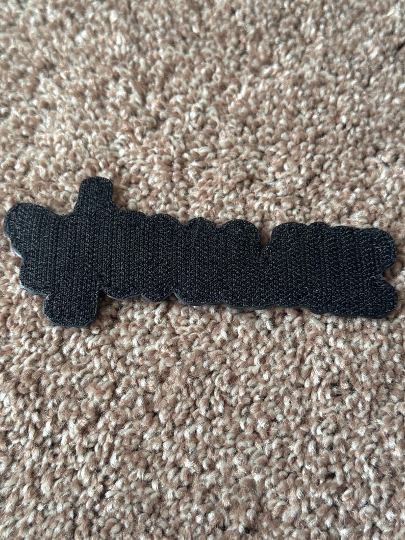 Pode incluir: Um patch preto com a palavra "dreamer" em fonte cursiva. O patch tem uma superf&iacute;cie texturizada e uma borda ondulada. O patch est&aacute; sobre um tapete castanho.