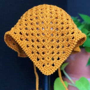 May include: A crocheted sun hat in a mustard yellow color. The hat has a square shape and is tied with long strings.