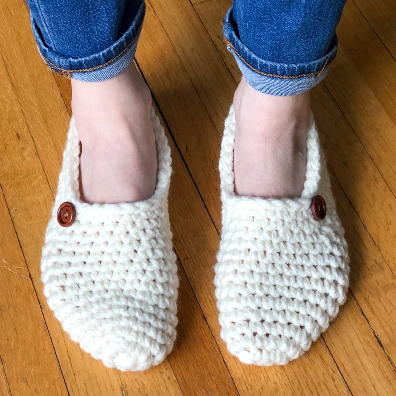 May include: A pair of white crocheted slippers with brown buttons. The slippers are worn by a person wearing blue jeans.