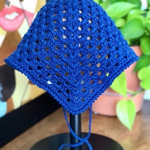 May include: A blue crocheted sun hat with a square pattern. The hat is displayed on a wooden stand with a black base.