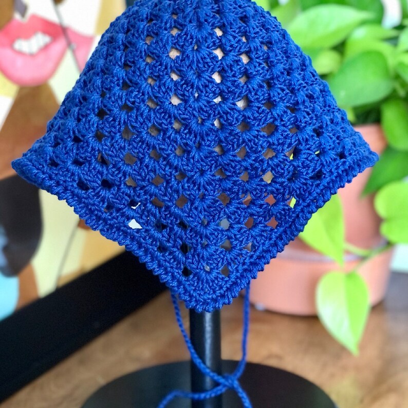 May include: A blue crocheted sun hat with a square pattern. The hat is displayed on a wooden stand with a black base.