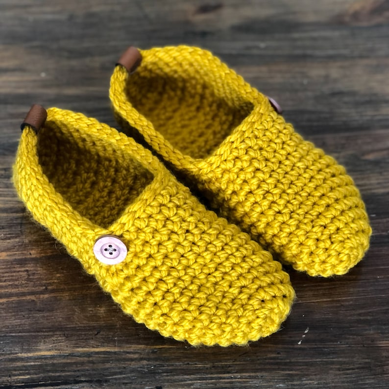 May include: A pair of yellow crocheted slippers with a pink button on each. The slippers have a brown leather trim around the opening.