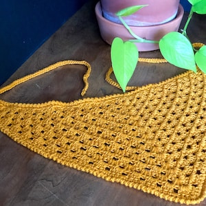 May include: A mustard yellow crocheted triangle scarf with a fringe edge. The scarf is lying on a wooden surface with a green plant in the background.