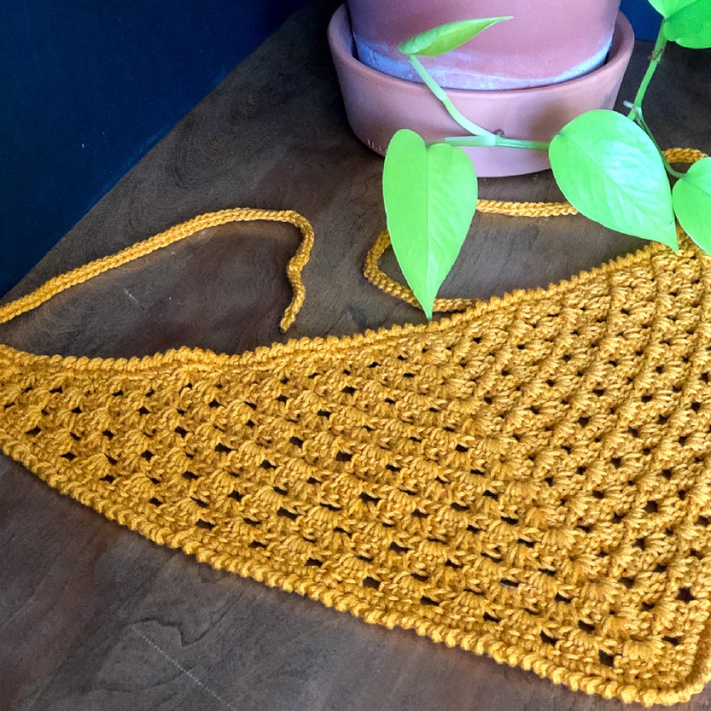 May include: A mustard yellow crocheted triangle scarf with a fringe edge. The scarf is lying on a wooden surface with a green plant in the background.