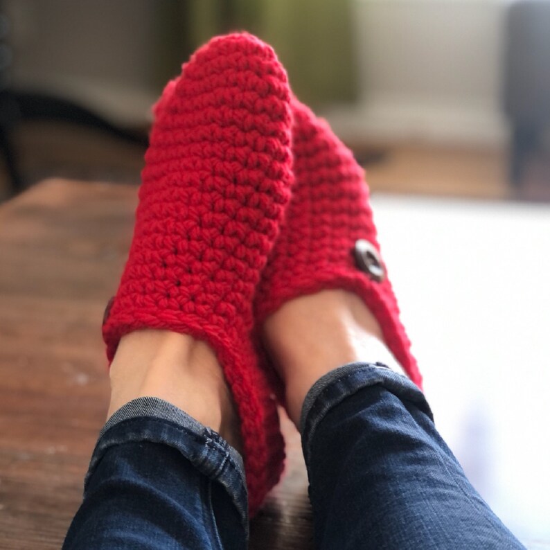 May include: Red crocheted slippers with a button detail. The slippers are worn on a person's feet.