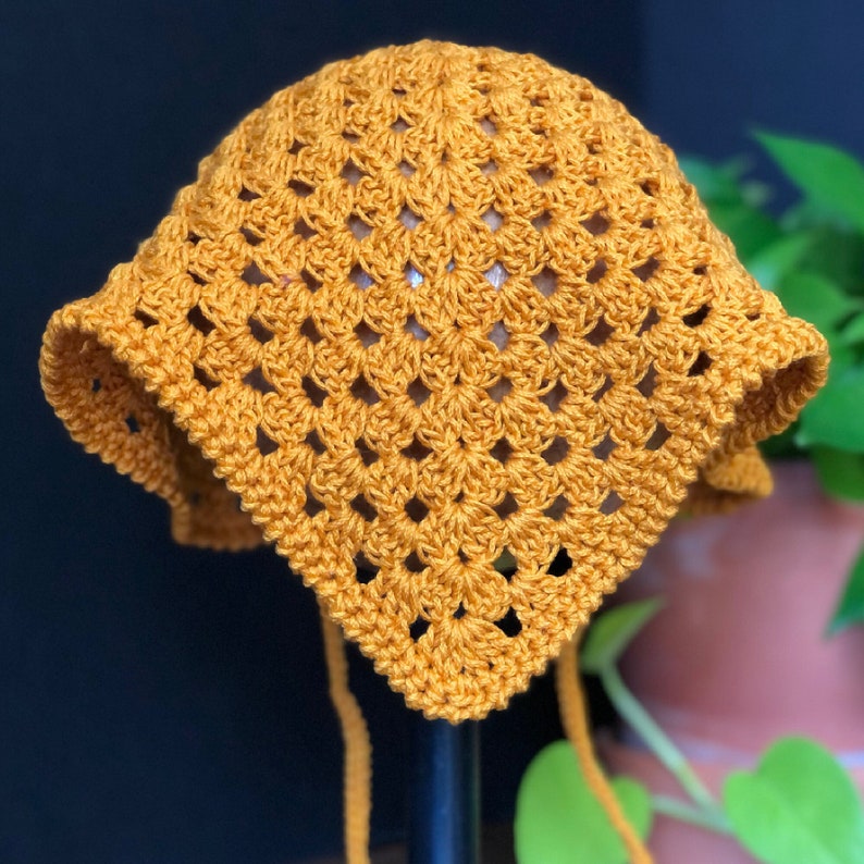 May include: A crocheted sun hat in a mustard yellow color. The hat has a square shape and is tied with long strings.
