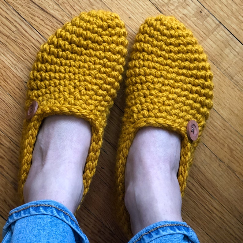 May include: A pair of yellow crocheted slippers with brown button accents. The slippers are worn on a person's feet.