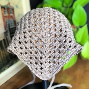 May include: A crocheted hat in a light beige color with a square stitch pattern. The hat is displayed on a mannequin head.