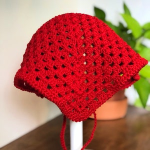 May include: A red crocheted hat with a square pattern and ties. The hat is displayed on a white stand.