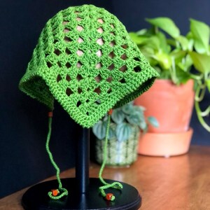 May include: Green crochet sun hat with a square pattern and orange beads on the ties. The hat is displayed on a black stand.