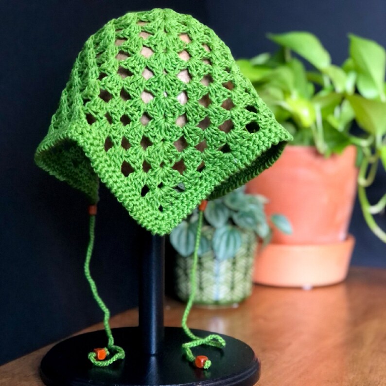 May include: Green crochet sun hat with a square pattern and orange beads on the ties. The hat is displayed on a black stand.