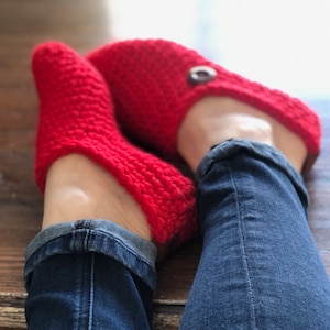 May include: A pair of red crocheted slippers with a button detail. The slippers are worn by a person wearing blue jeans.