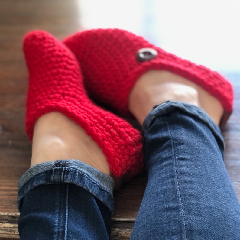 May include: A pair of red crocheted slippers with a button detail. The slippers are worn by a person wearing blue jeans.