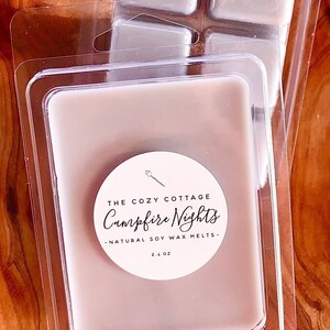 May include: Four wax melts in a clear plastic container. The wax melts are a light brown color. The container has a white label with the text "The Cozy Cottage Campfire Nights - Natural Soy Wax Melts - 2.4 oz"
