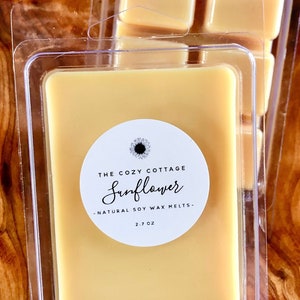 May include: A clear plastic container with four wax melts. The wax melts are a light yellow color. The container has a white label that says "The Cozy Cottage Sunflower - Natural Soy Wax Melts - 2.7 oz."