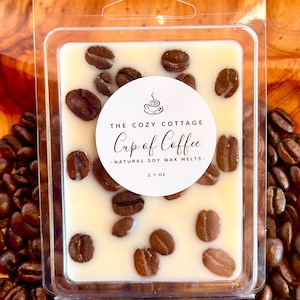 May include: A clear plastic container holds a creamy-colored wax melt, adorned with coffee beans. The label reads "The Cozy Cottage Cup of Coffee - Natural Soy Wax Melts - 2.7 oz." The container rests on a bed of coffee beans, with a wooden background.