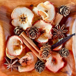 May include: A wooden bowl filled with dried apple slices, cinnamon sticks, star anise, pine cones, and vanilla beans. The bowl is on a wooden surface.