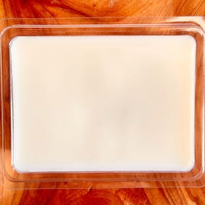 Puede incluir: Un derrite de cera rectangular, blanquecino, en un recipiente de pl&aacute;stico transparente. La cera tiene una superficie lisa y est&aacute; rodeada por un marco de pl&aacute;stico transparente. El recipiente est&aacute; sobre una superficie de madera.