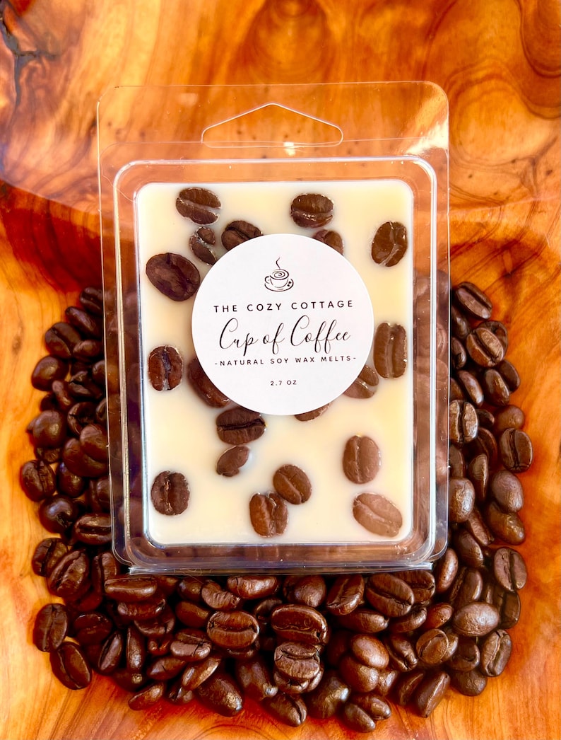 May include: A clear plastic container holds a creamy-colored wax melt, adorned with coffee beans. The label reads "The Cozy Cottage Cup of Coffee - Natural Soy Wax Melts - 2.7 oz." The container rests on a bed of coffee beans, with a wooden background.