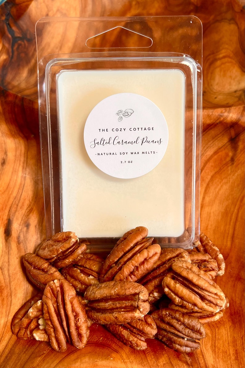 May include: A clear plastic container holds a creamy-colored soy wax melt. The label reads "The Cozy Cottage Salted Caramel Pecans - Natural Soy Wax Melts - 2.7 oz." Several pecan halves are scattered around the container.