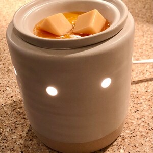 May include: A white ceramic wax warmer with a beige base. The warmer has a round top with a well for melting wax. Two square wax melts are in the well, surrounded by melted wax.
