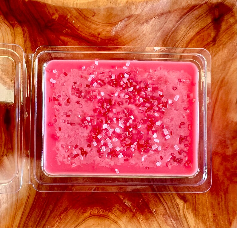 May include: A pink wax melt with white sprinkles in a clear plastic container.