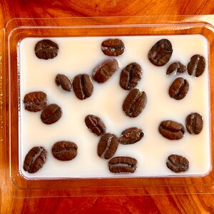 May include: A rectangular, clear plastic container holding a creamy white substance with several dark brown coffee beans embedded within. The beans are scattered across the surface, creating a visually appealing arrangement. The container is on a wooden surface.
