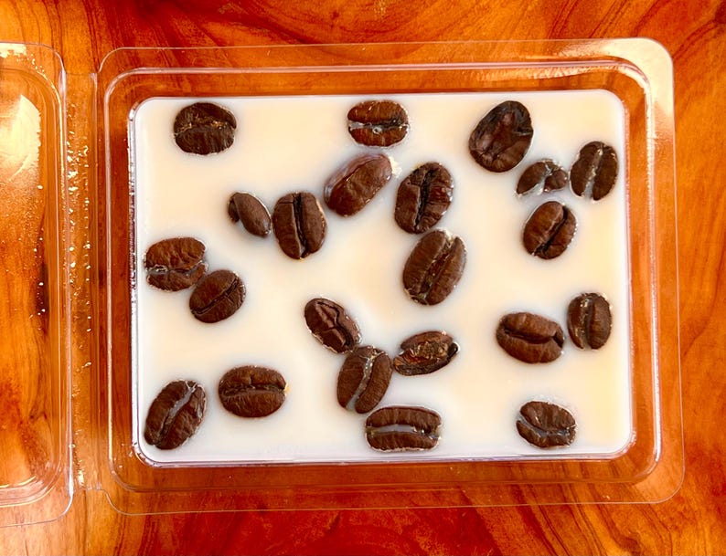 May include: A rectangular, clear plastic container holding a creamy white substance with several dark brown coffee beans embedded within. The beans are scattered across the surface, creating a visually appealing arrangement. The container is on a wooden surface.