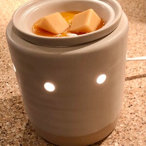 May include: A white ceramic wax warmer with two wax melts inside. The warmer has a round top with a well for the melts and a base with holes for light to shine through. The wax melts are a light yellow color and are melting in the well.