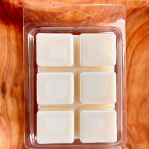 May include: A clear plastic clamshell package containing six off-white wax melt cubes. The cubes are arranged in two rows of three. The background is a wooden surface.