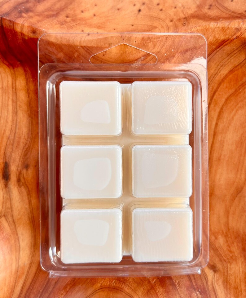 May include: A clear plastic clamshell package containing six off-white wax melt cubes. The cubes are arranged in two rows of three. The background is a wooden surface.