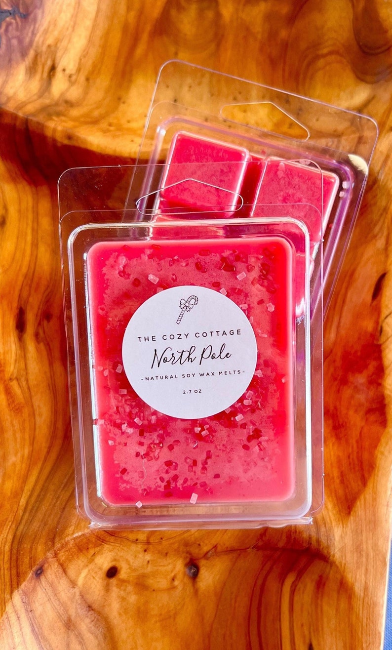 May include: A red wax melt in a clear plastic container with a white label that reads "The Cozy Cottage North Pole - Natural Soy Wax Melts - 2.7 oz".