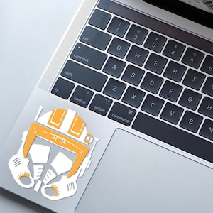 May include: A white and orange sticker of a Star Wars clone trooper helmet on a silver laptop.