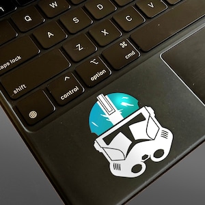 May include: A white and blue sticker of a Star Wars clone trooper helmet on a black laptop keyboard.
