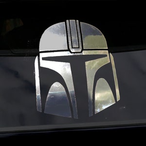 May include: A silver Mandalorian helmet sticker, featuring the iconic design from the Star Wars series. The sticker is applied to a dark surface, likely a car window, and reflects light, highlighting the helmet's shape and details. A fan-made item.