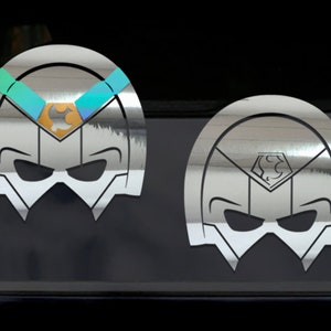 May include: Two silver-colored mask-shaped decals. One has a gold and teal accent across the top, while the other has a silver emblem. The decals are on a dark surface. The masks have a stylized design.