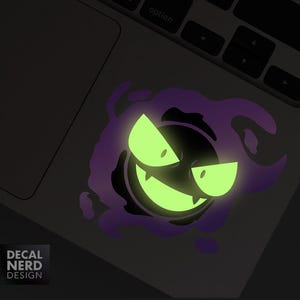 May include: A glowing sticker featuring a cartoon ghost with a mischievous grin and glowing green eyes. The ghost is surrounded by a purple aura. The sticker is on a laptop.