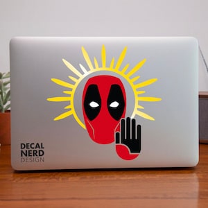 May include: A laptop computer with a red and black Deadpool sticker on the lid. The sticker features Deadpool's mask with a halo of yellow sun rays and a black hand with a raised index finger. The text "DECAL NERD DESIGN" is printed on the bottom of the laptop.
