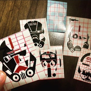May include: Five black and red vinyl decals on white sheets with a grid pattern. The decals are of different Star Wars characters, including a Stormtrooper, a Mandalorian, and a clone trooper.